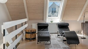 Una habitación con techo inclinado, una ventana con vista a una casa, dos sillas negras, un taburete de madera y un radiador.