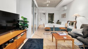 Camera d'albergo moderna con televisione a schermo piatto, mobile TV in legno, una pianta grande, un letto con testiera verde, un divano, un tavolino da caffè e una lampada.