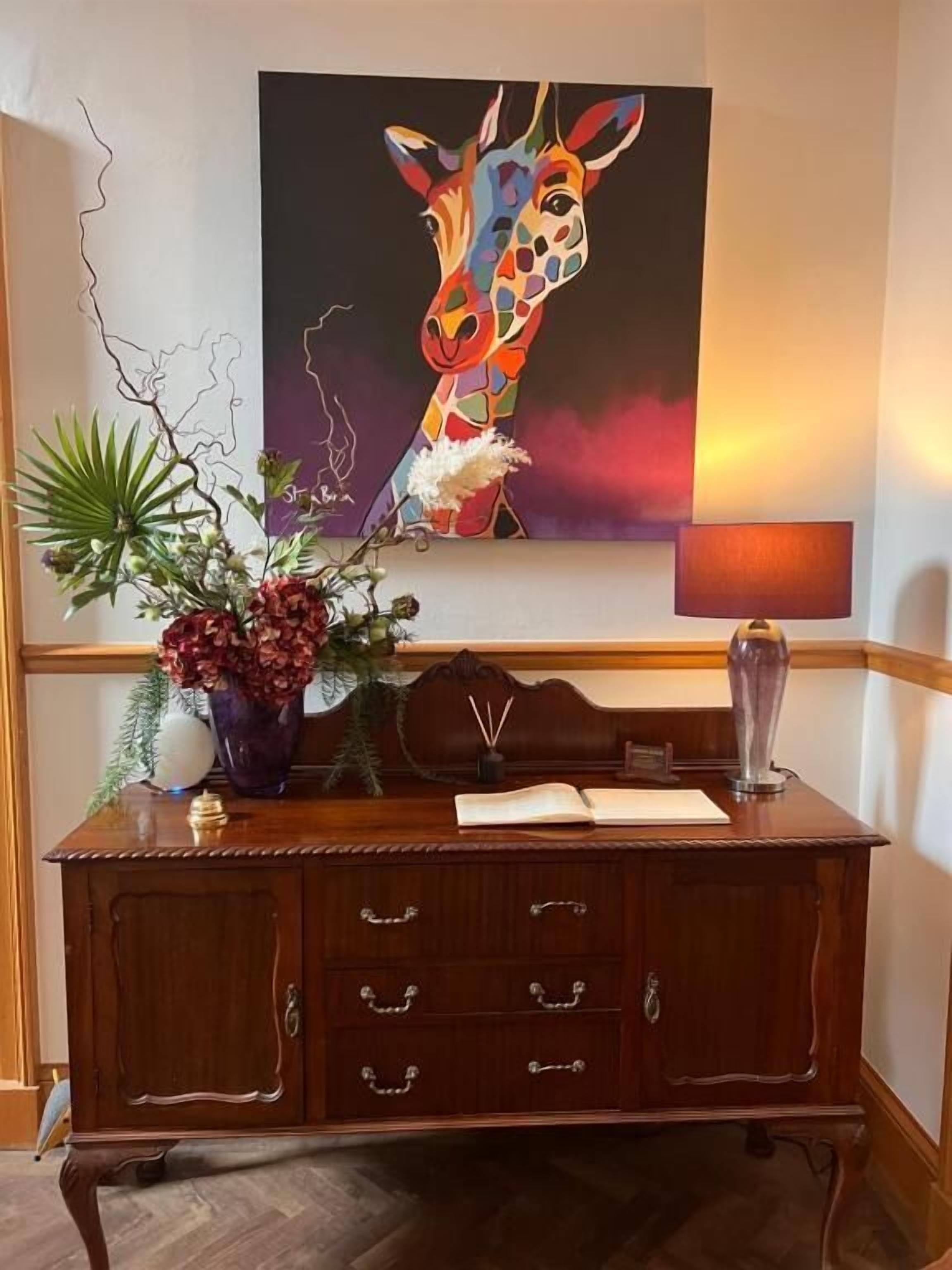 A wooden desk with a vase of flowers, a lamp, and a painting of a giraffe on the wall.