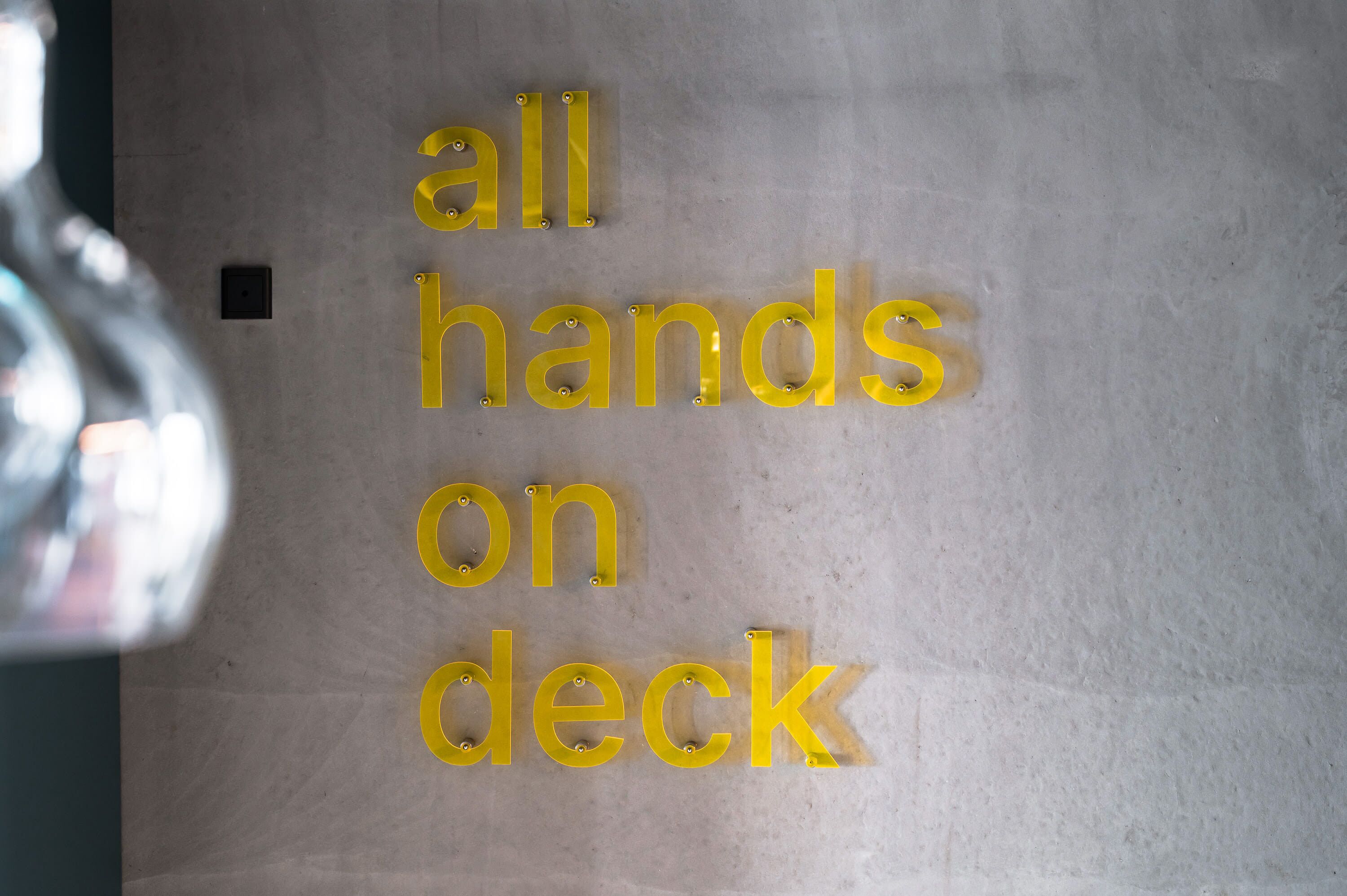 A sign with the text "all hands on deck" mounted on a wall.