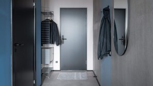 Um corredor com uma porta fechada, uma estante de metal com roupas penduradas e um espelho na parede.