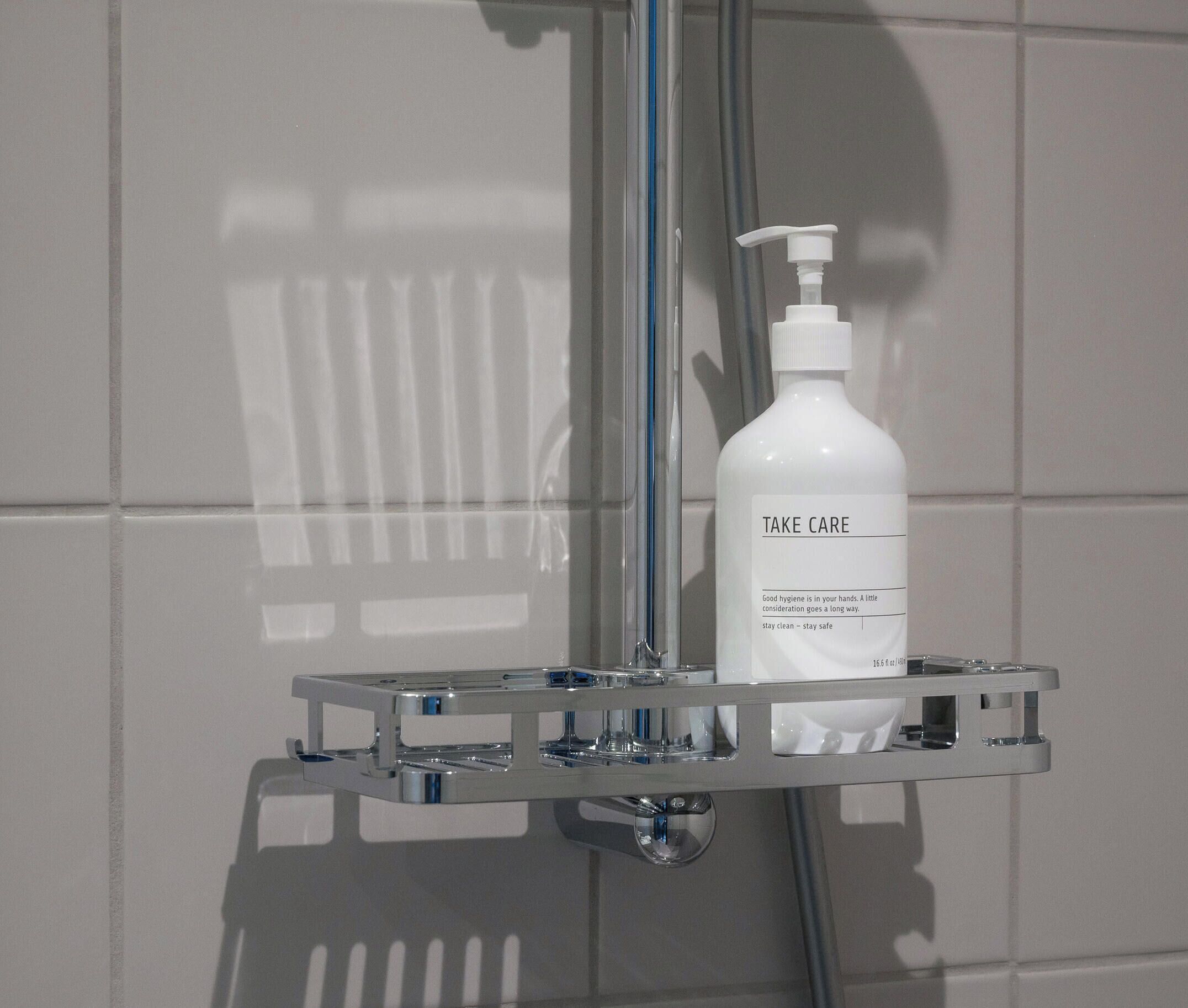A bathroom with a white tiled wall, a chrome shower caddy holding a bottle of 'Take Care' hand soap, and a showerhead.