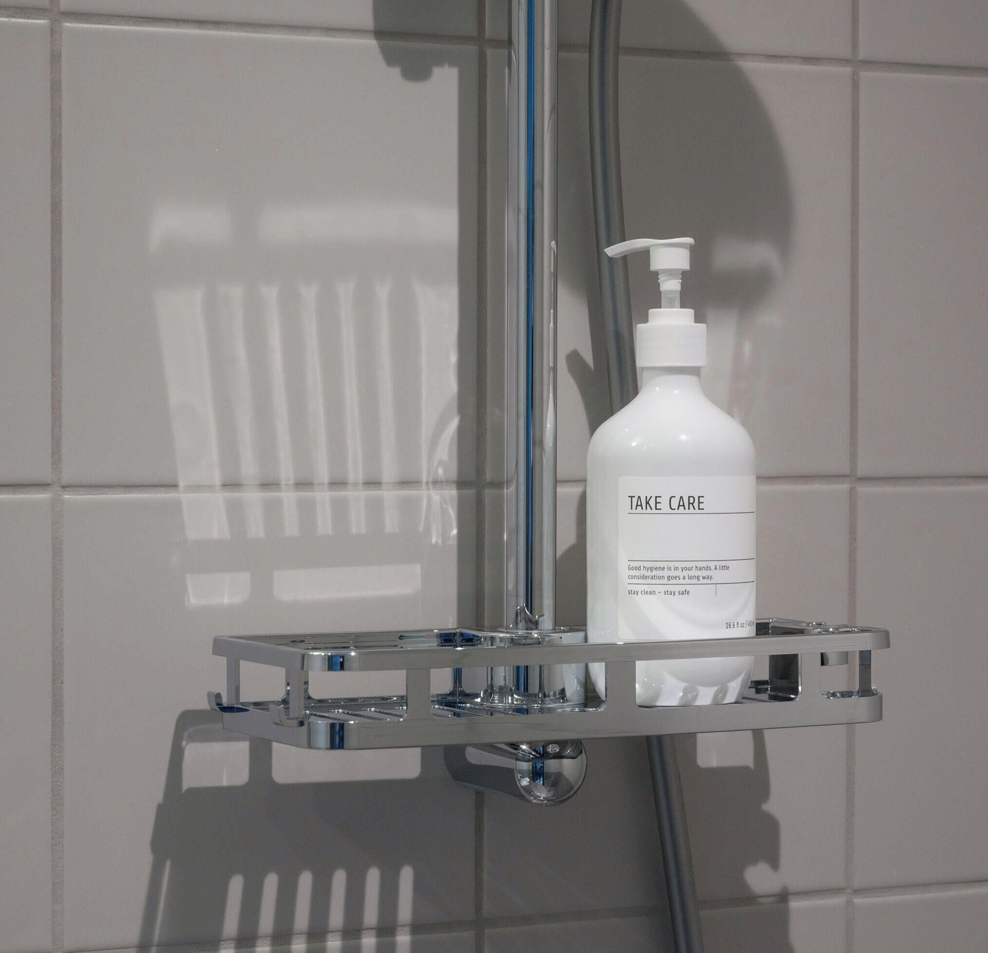 Une salle de bain avec un mur recouvert de carreaux blancs, un porte-savon chromé contenant un flacon de savon liquide « Take Care », et une pomme de douche.