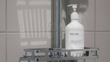 Une salle de bain avec un mur recouvert de carreaux blancs, un porte-savon chromé contenant un flacon de savon liquide « Take Care », et une pomme de douche.