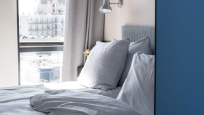 Una cama bien hecha con sábanas blancas en una habitación con un ventanal grande, una lámpara de pared y vistas a edificios.