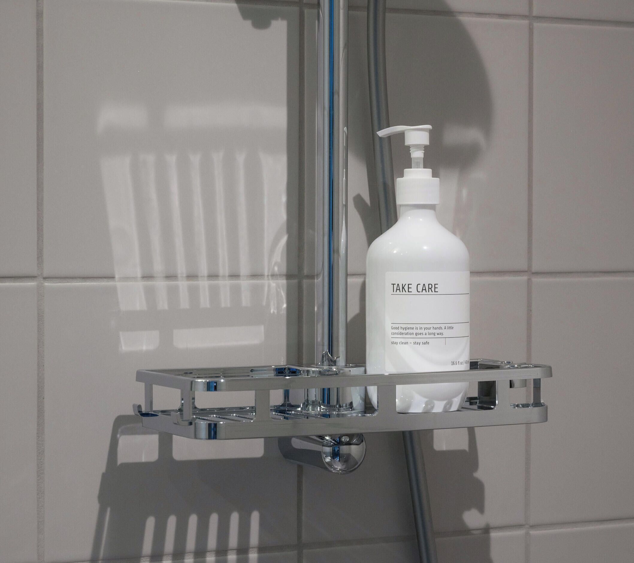 Een badkamer met een witte betegelde muur, een chroom douchemandje met een fles 'Take Care' handzeep, en een douchekop.