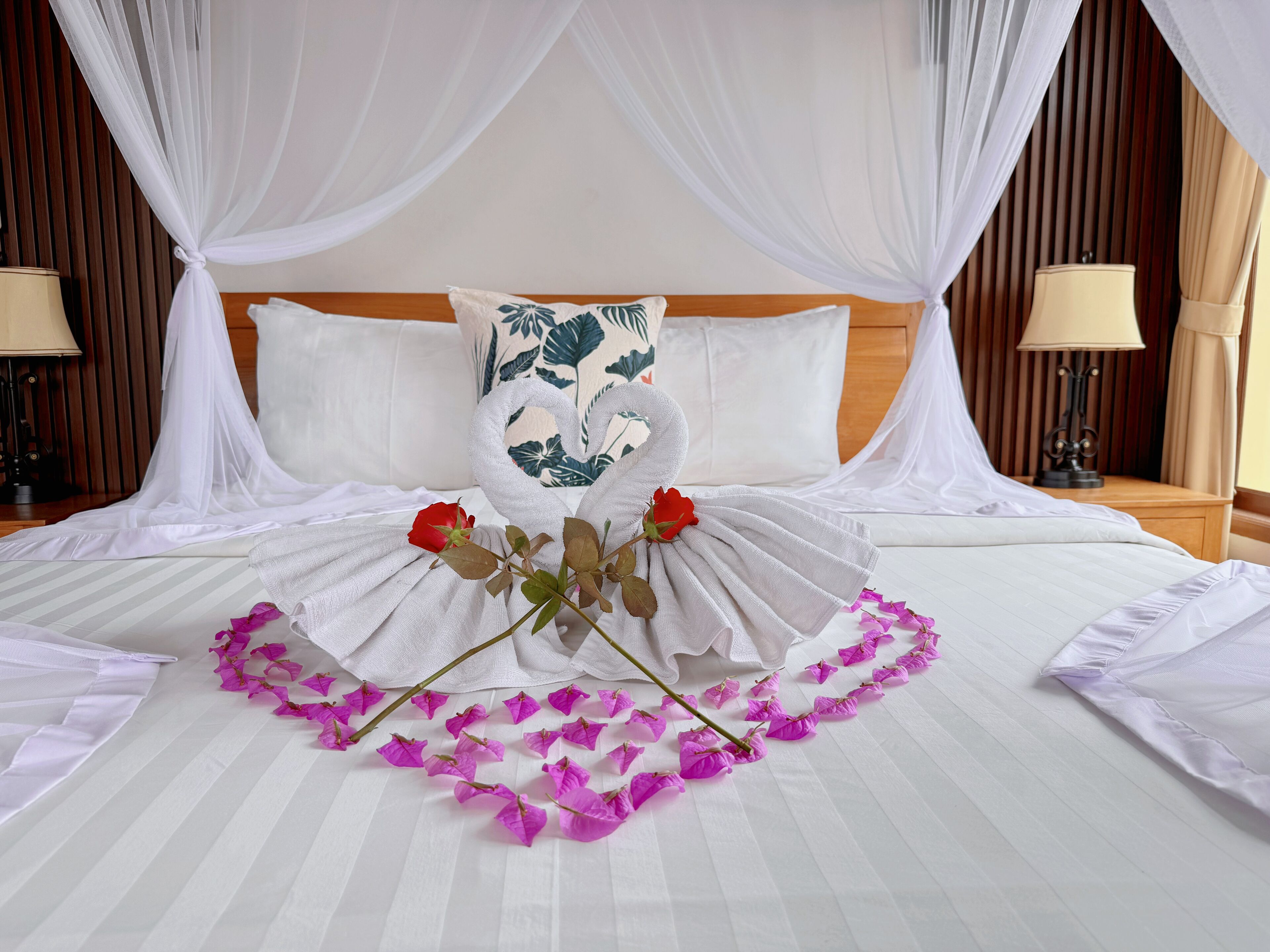 Una cama bien hecha con una colcha blanca, un cojín decorativo con estampado de hojas verdes y un ramo de flores rojas y púrpuras.