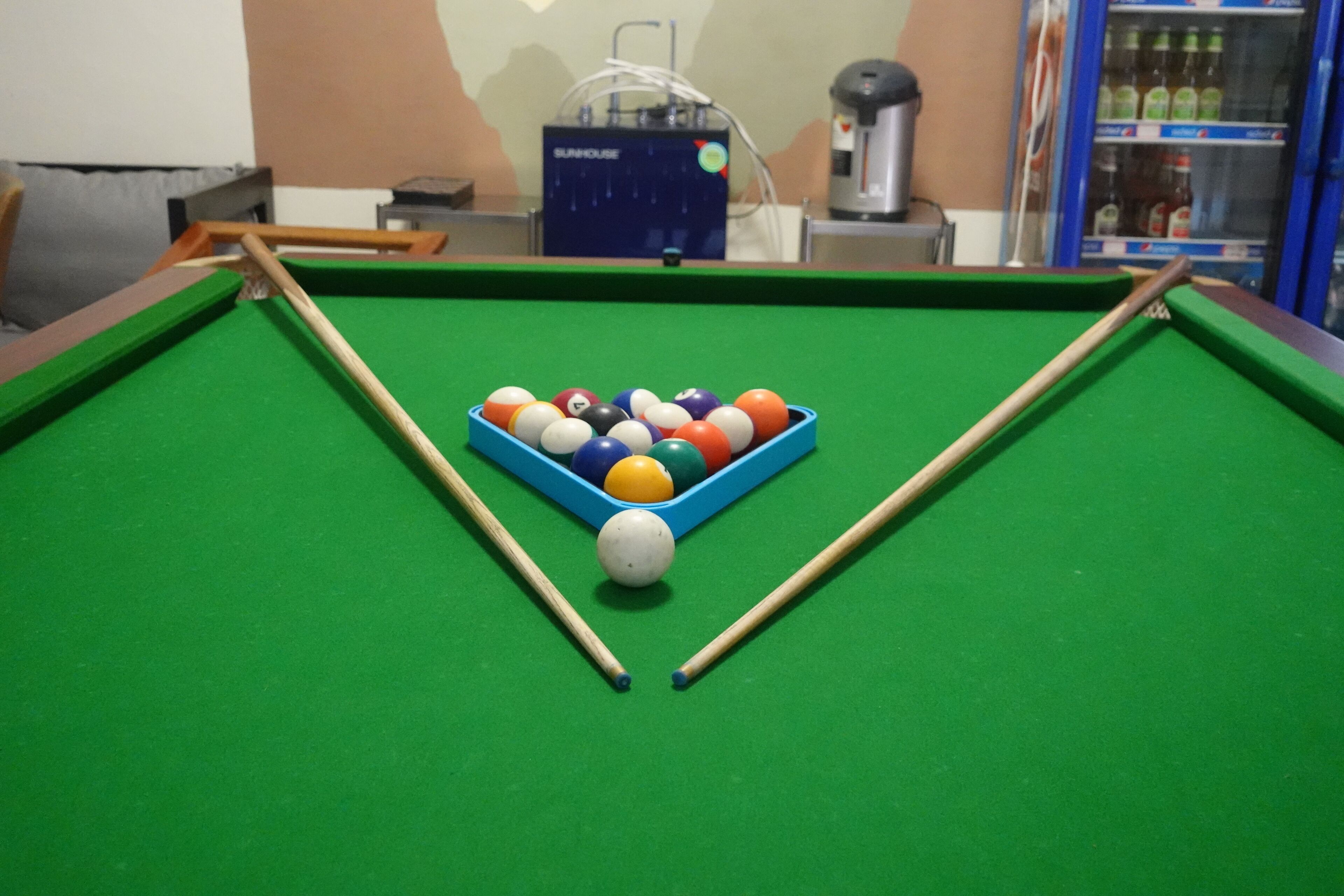 A pool table with a blue corner rack holding various colored balls, a cue stick, and a white ball in the center.