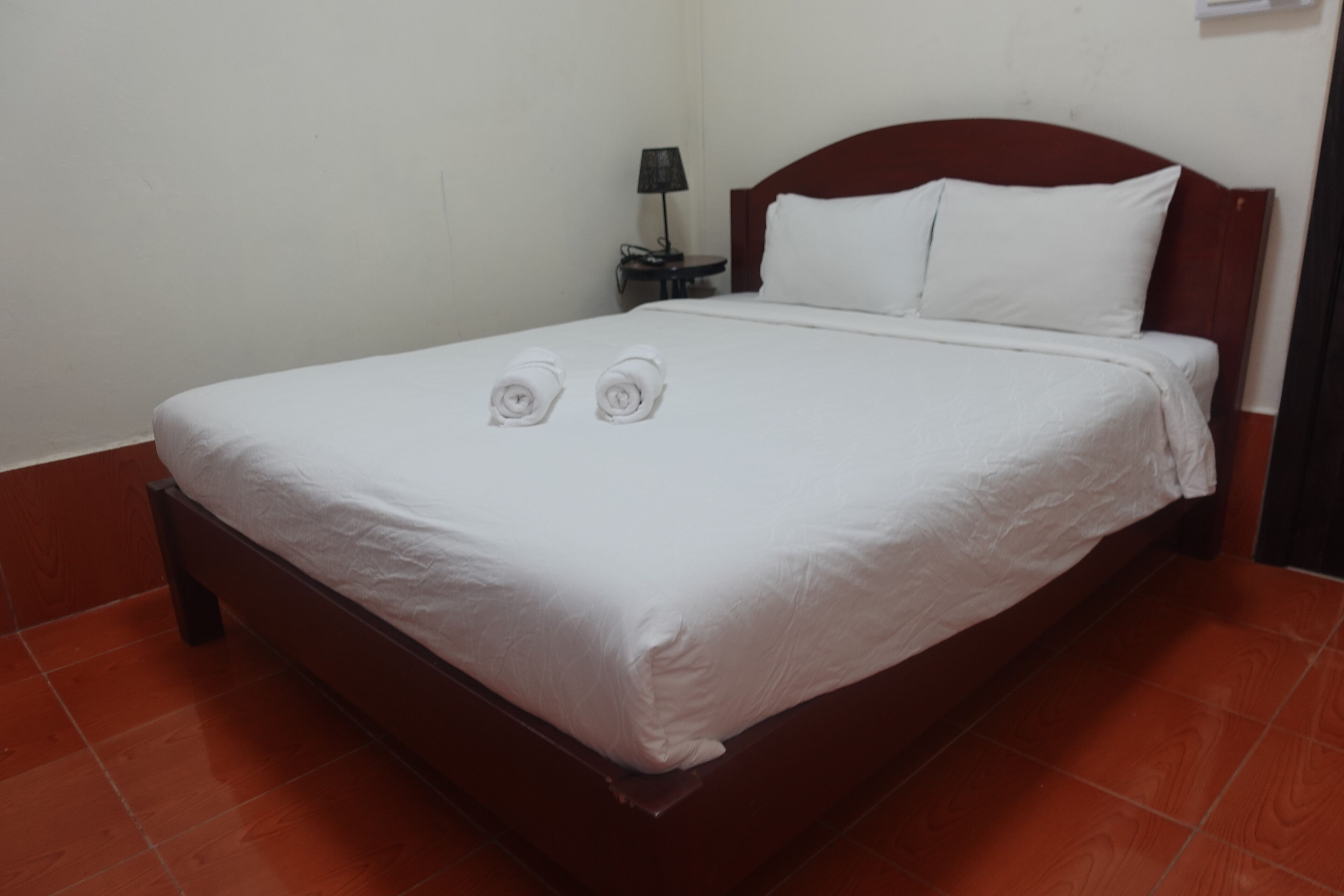 A neatly made bed with white linens and two rolled towels, a wooden headboard, a bedside table with a lamp, and a red-tiled floor.