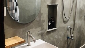 Une salle de bain avec un lavabo blanc, un miroir rond, une douchette à main et une petite étagère contenant des articles de toilette.