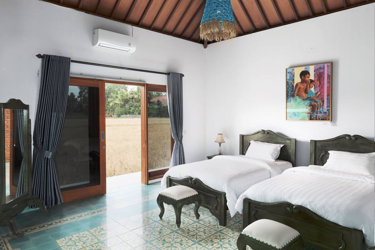 Una habitación con dos camas, un ventanal con cortinas, un ventilador de techo y un cuadro en la pared.