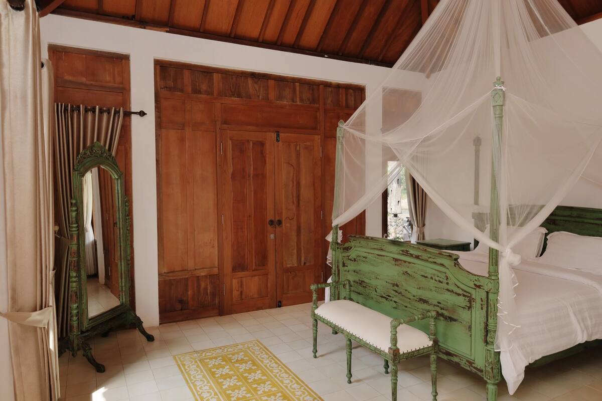 Un dormitorio con una cama con dosel, un sillón antiguo verde, un armario de madera y un espejo.
