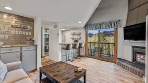 A modern living room with a wooden coffee table, a sofa, and a kitchen area with bar stools. - Free Ski Locker@Resort Base-Views-Pool-HotTub-Grill-Deck+Walk to Mtn/Trails/Eat/Drink/Free Town Bus (Steamboat Springs)