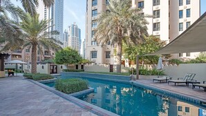 A swimming pool with a tiled deck, surrounded by palm trees and a building with multiple floors in the background. - Stylish 2BR with Balcony Near Dubai Mall & Burj Khalifa (Oldtown Dubai)