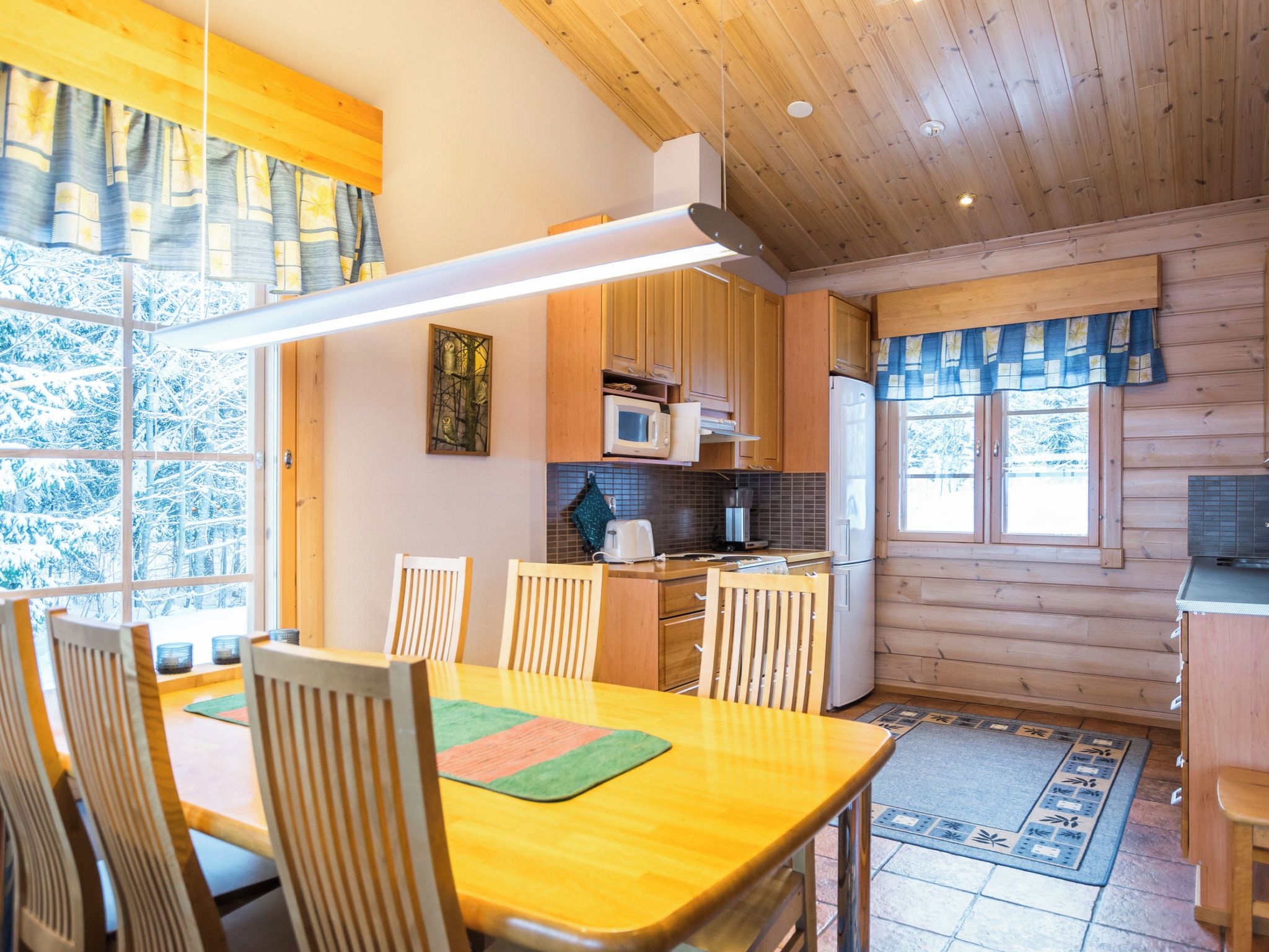 Une cuisine chaleureuse, aménagée dans le style d’un chalet, avec des meubles en bois, une table à manger avec des chaises et une grande fenêtre agrémentée de rideaux.