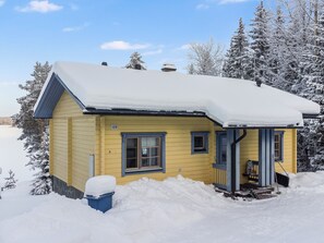 Une maison jaune, dont le toit est recouvert de neige, entourée d’arbres également couverts de neige.