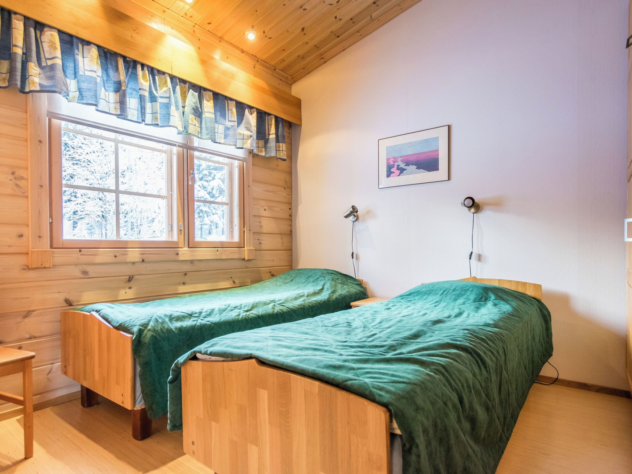 A room with two wooden beds, a window with curtains, a painting on the wall, and a lamp.