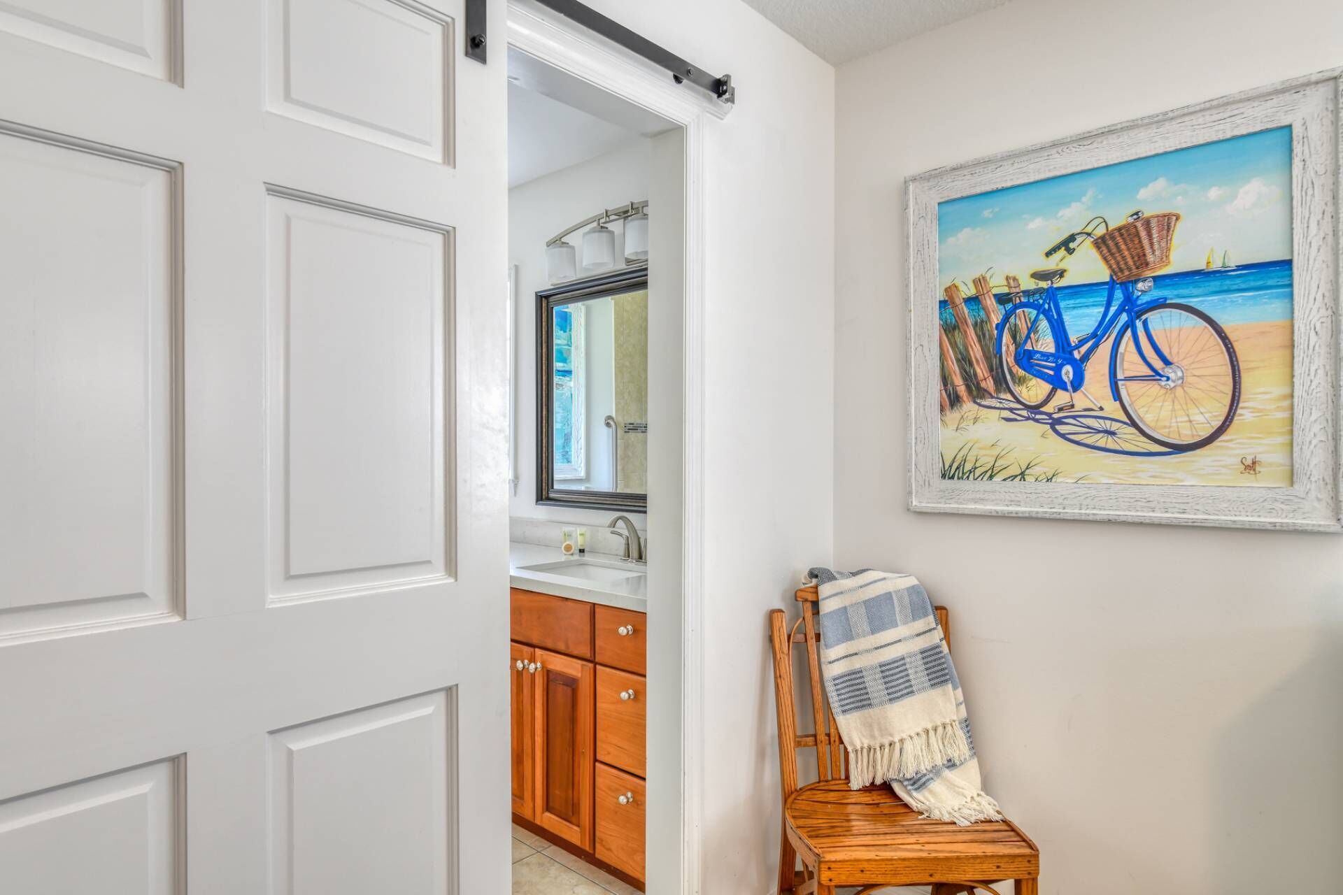 A bathroom with a white door, a wooden cabinet, a mirror, and a framed painting of a blue bicycle on a beach.