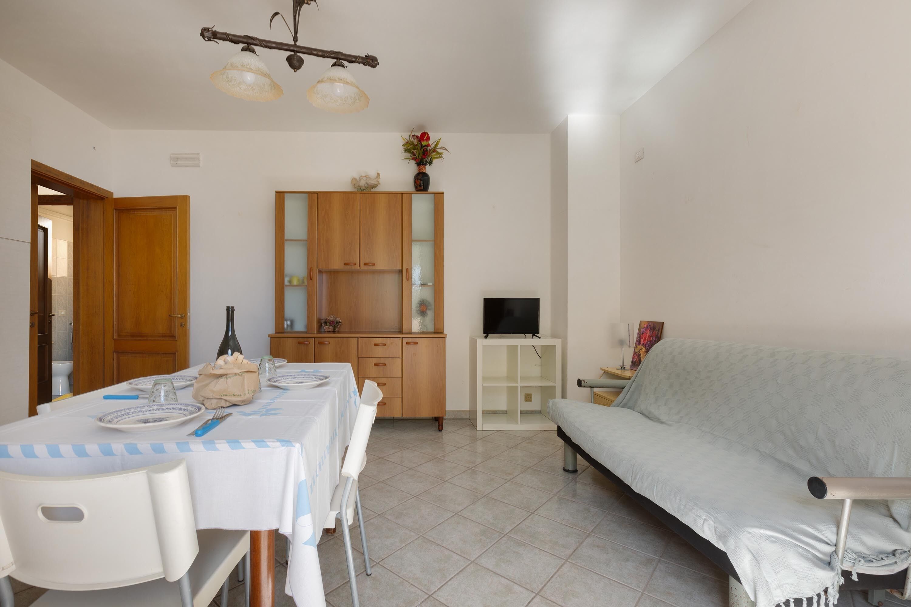 A room with a dining table set for two, a sofa, a cabinet with glass doors, and a television on a stand.
