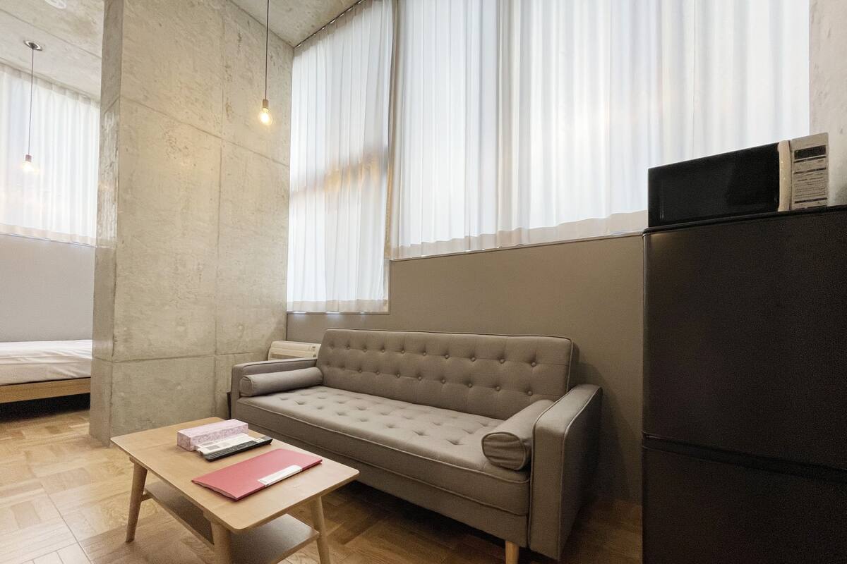 Quarto moderno com sofá, uma pequena mesa, uma cama, frigorífico e janelas amplas com cortinas leves.