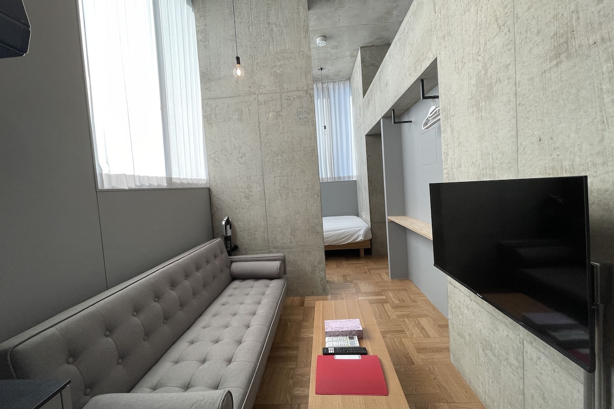 Um espaço de estar moderno e compacto, com um sofá cinza, uma mesa de centro de madeira, uma televisão e uma cama ao fundo.