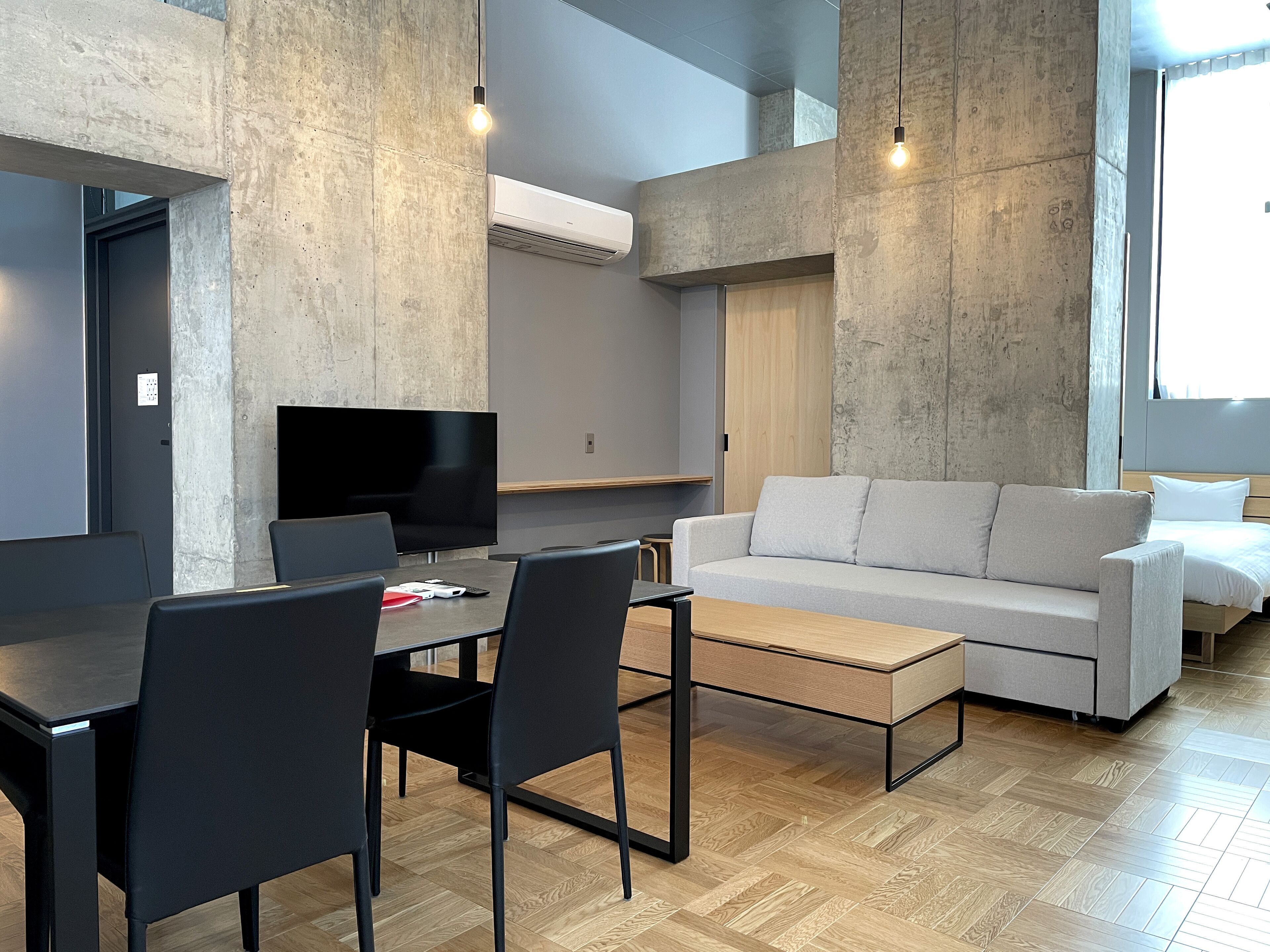 A modern living room with a grey sofa, a coffee table, and a TV. There are two black chairs and a dining table. The room has a concrete accent wall and a wooden floor.