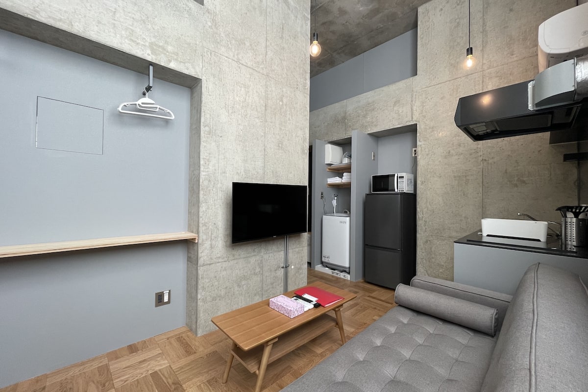 Um espaço de estar compacto com televisão de tela plana na parede, uma pequena mesa de madeira, um sofá cinza, um espaço de cozinha com micro-ondas e geladeira, e uma luminária pendente.