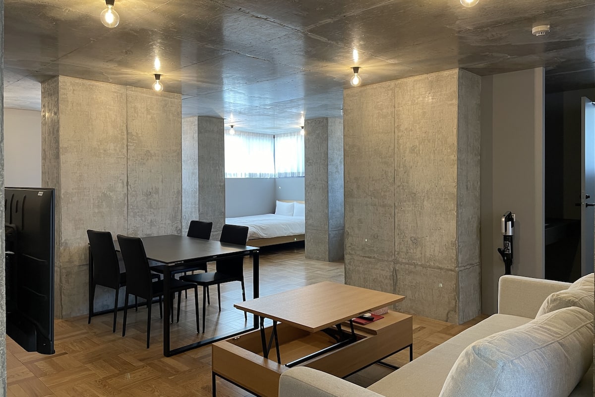 Quarto moderno em estilo industrial, com mesa de jantar de madeira, cadeiras pretas, sofá e cama com roupa de cama branca.