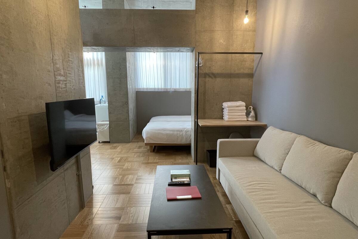 Quarto moderno com televisão de tela plana, um sofá, uma pequena mesa com um livro e uma cama com vista para a janela.