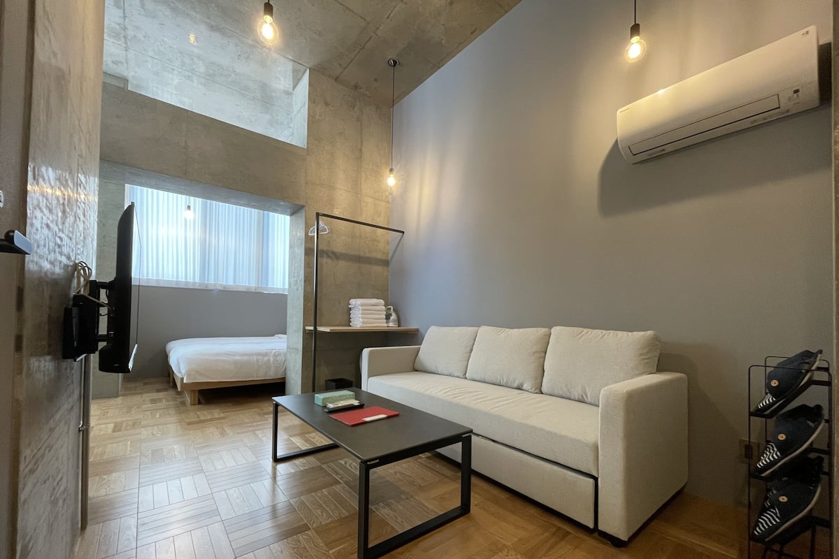 Quarto de hotel moderno com sofá, mesa pequena, cama, sapateira e aparelho de ar condicionado.