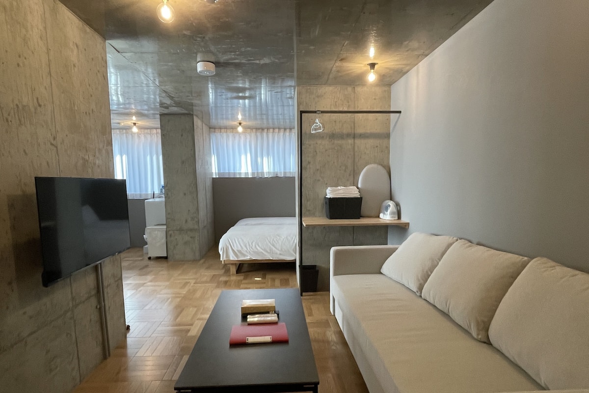 Quarto moderno e minimalista com televisão de tela plana, sofá, cama e uma pequena mesa com livros.