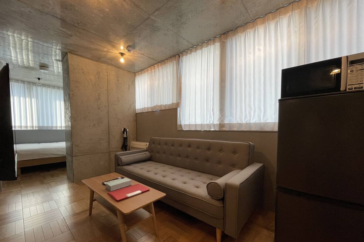 Quarto compacto com sofá, uma pequena mesa, geladeira e janela com cortinas.