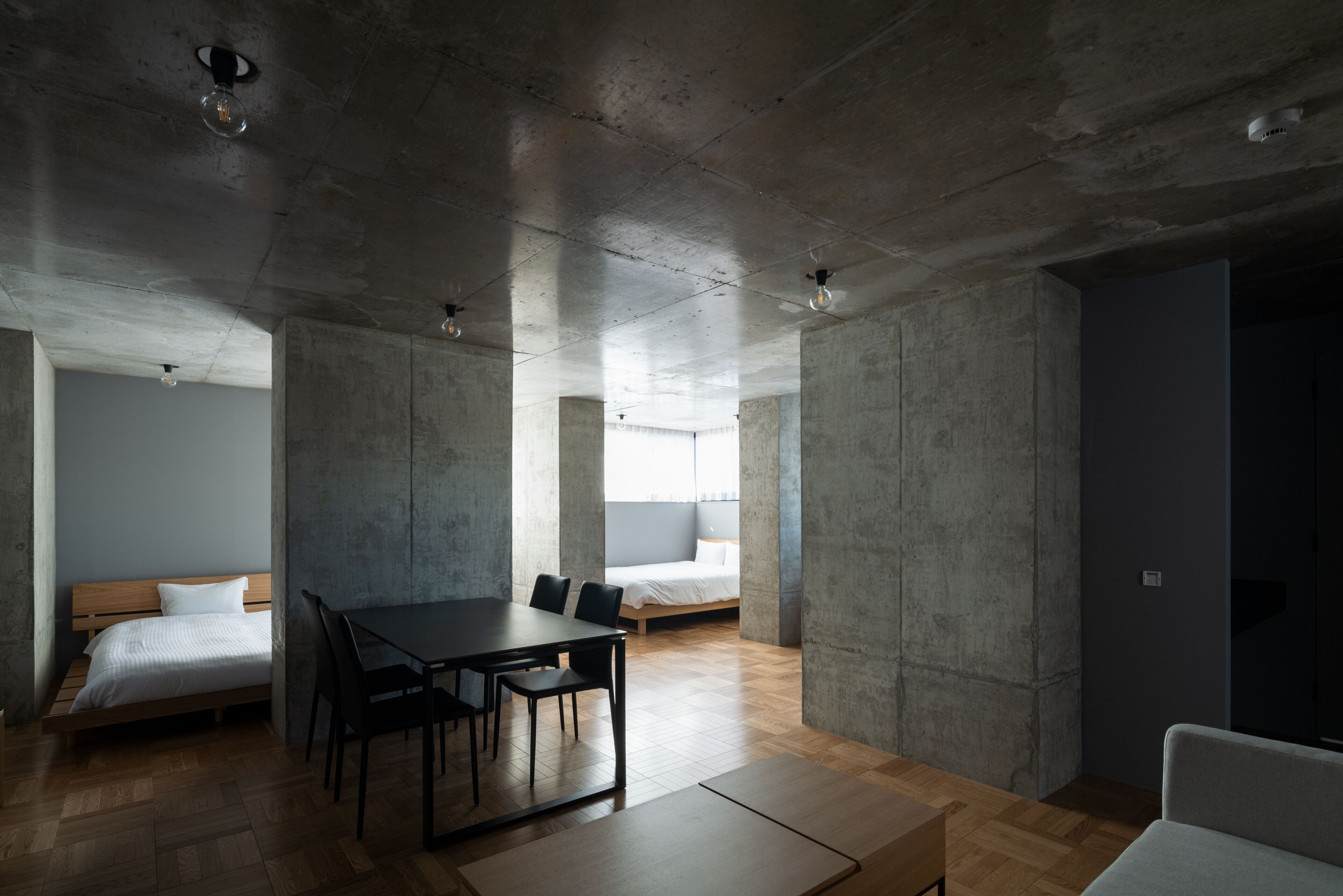 Une chambre d’hôtel moderne avec un plancher en bois, un lit avec une literie blanche, une table à manger avec des chaises et une grande fenêtre laissant entrer la lumière naturelle.