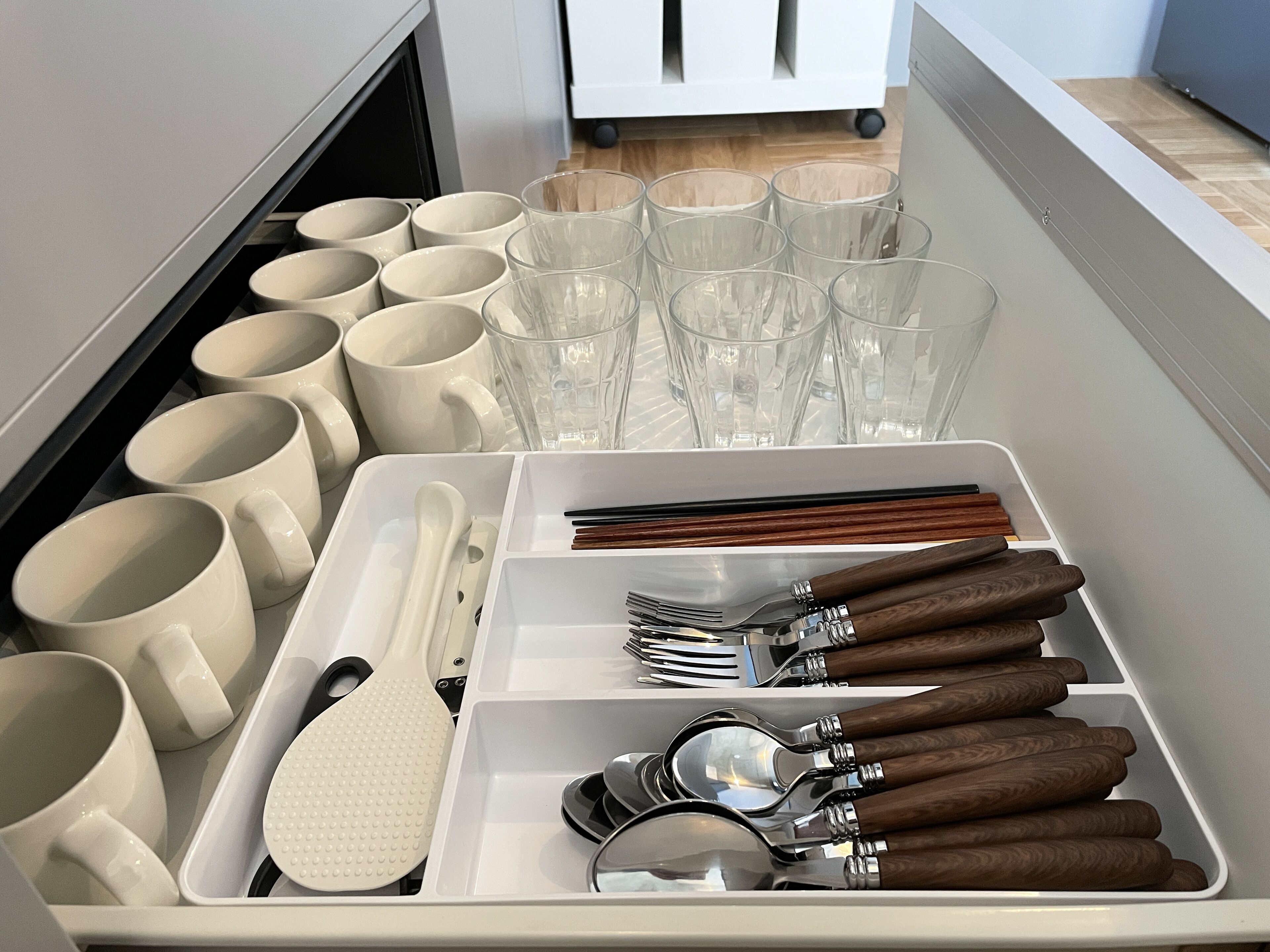 Un tiroir contenant des ustensiles de cuisine rangés avec soin, notamment des tasses, des verres, des cuillères, des fourchettes et des couteaux.