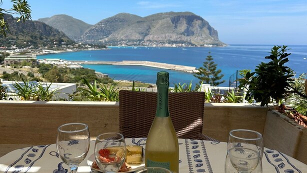 A table set with wine glasses, a bottle of wine, and a plate of food, overlooking a coastal landscape with mountains and the sea. - La Scalinatella in Mondello (Palermo)