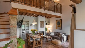 A cozy living room with a wooden dining table, a sofa, and a fireplace. There is a balcony with a wooden railing above the dining area.