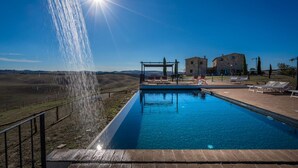 A swimming pool with a water feature, a pergola, and stone buildings in the background.