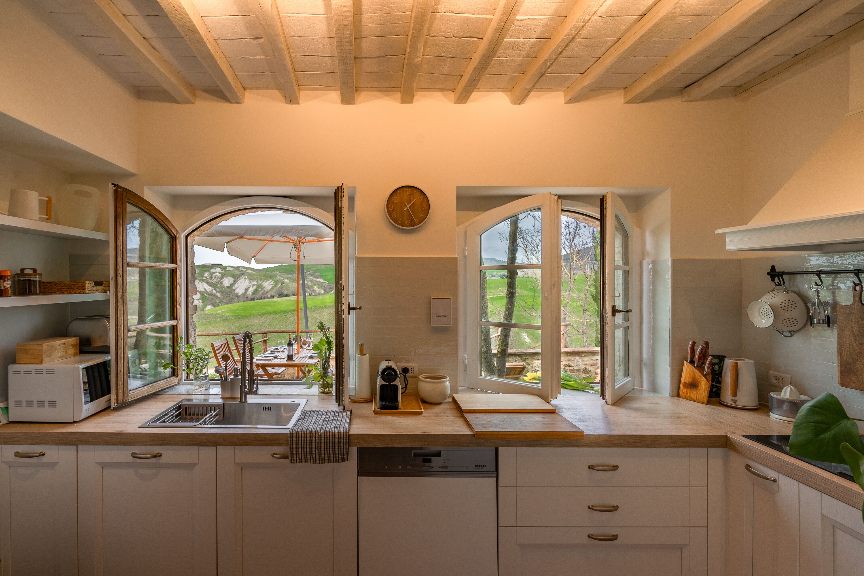 Cozinha com vista para o exterior, armários de madeira, pia e relógio na parede.