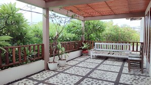 A covered outdoor area with a tiled floor, potted plants, a white bench, and a wooden fence. - Ananda's Sanur (Denpasar)