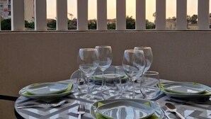Et rundt spisebord med tallerkener, glas og bestik dækket til seks personer. Bordet står på en balkon med udsigt til bygninger og en klar himmel.