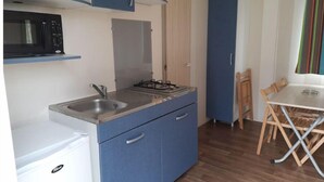 A compact kitchen with a microwave, sink, and stove. There is a small refrigerator, a dining table with chairs, and a cabinet with a door. - Mobil-home C007 (Boofzheim)