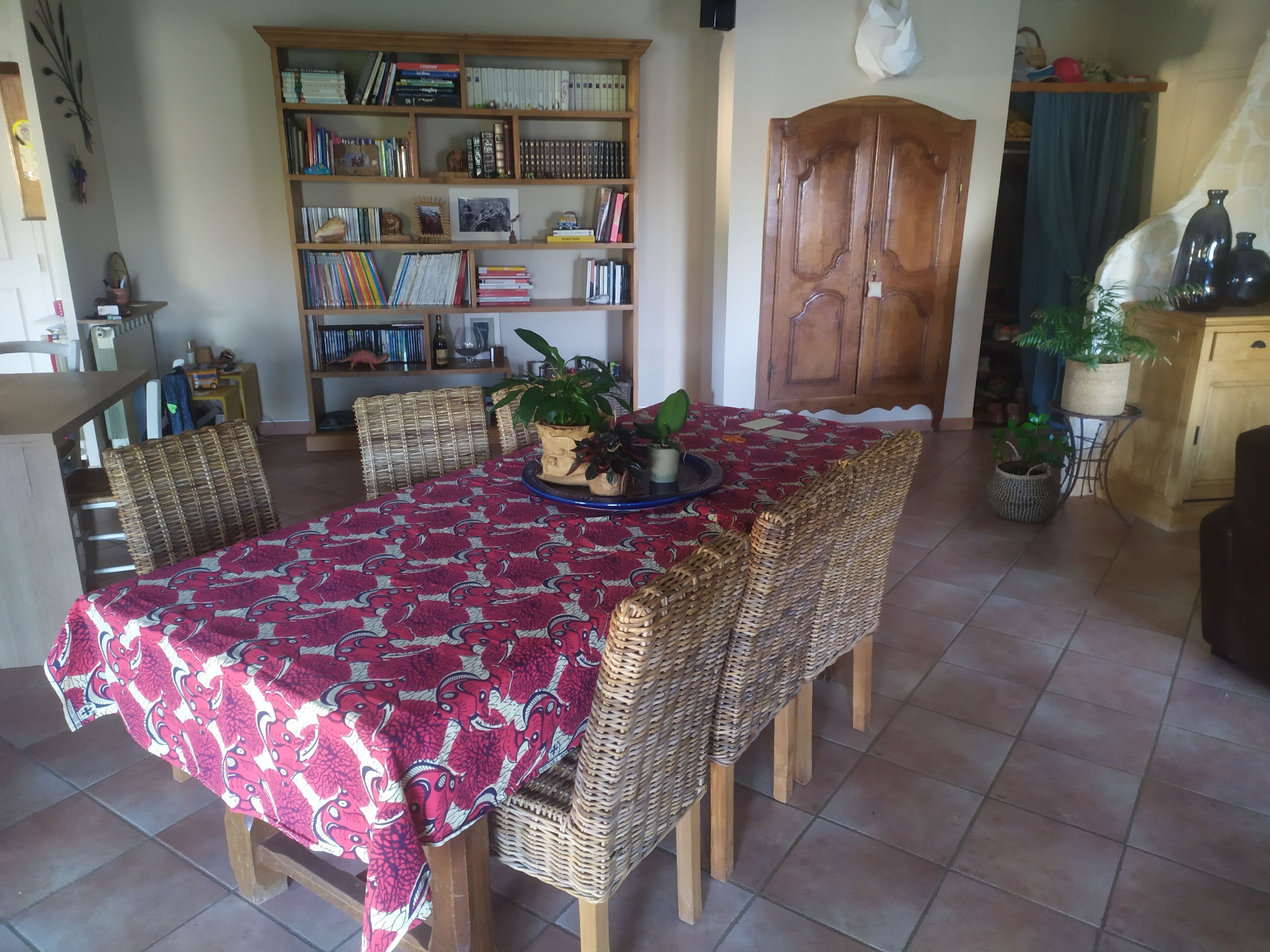 Un coin repas avec une table recouverte d’une nappe à motifs, entourée de chaises en osier. Une bibliothèque remplie de livres et d’objets décoratifs se trouve à l’arrière-plan.