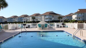 A swimming pool with a shallow water diving area, surrounded by a white fence and a row of apartment buildings in the background.