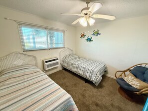 A bedroom with two single beds, a ceiling fan, a window with blinds, and a wall-mounted air conditioner. - Charming 2-bedroom condo offering serene living with picturesque pond vistas, with a lovely pool! (North Myrtle Beach)