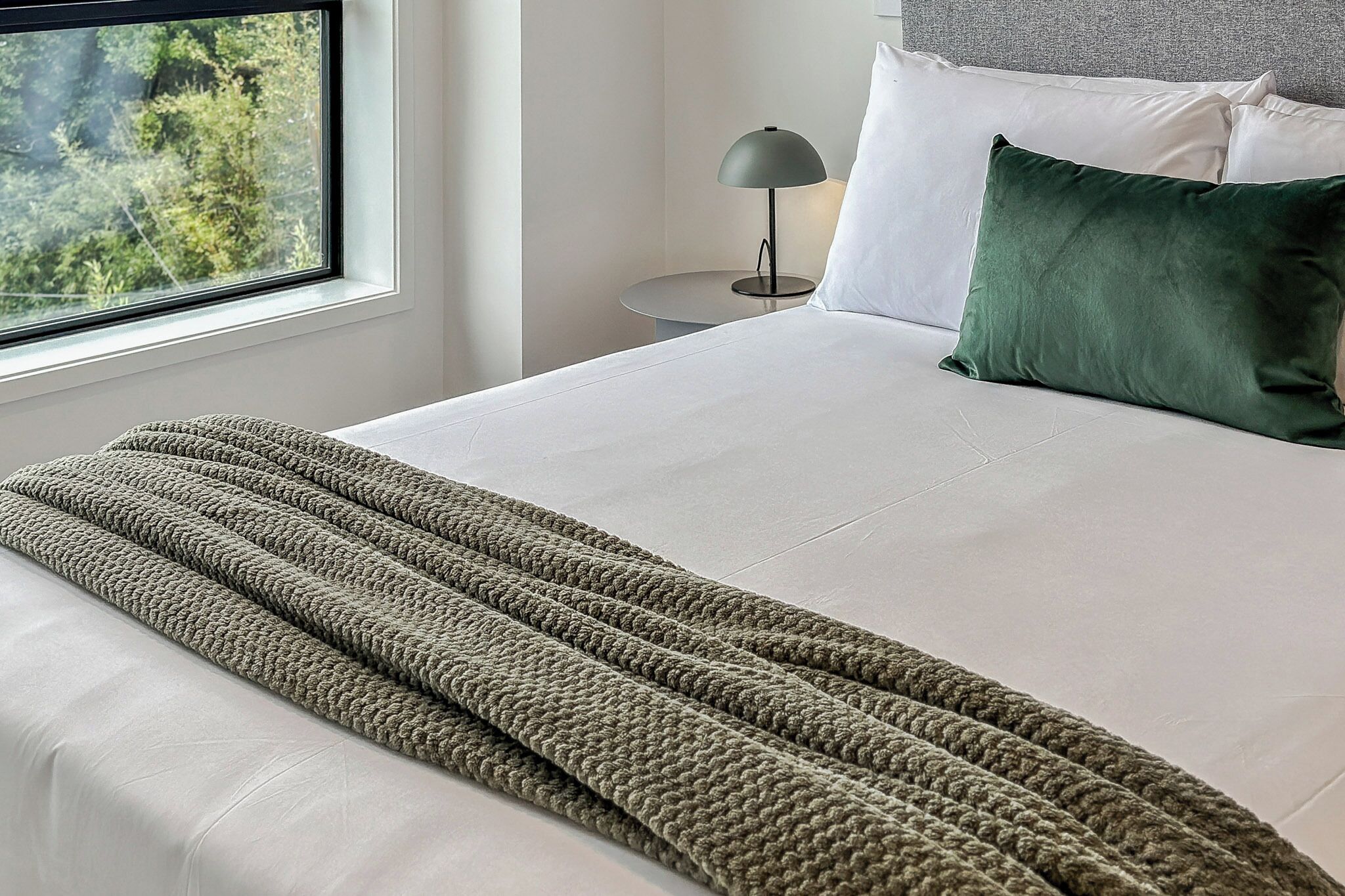 Una cama bien hecha con un plaid verde de punto, almohadas blancas y una lámpara gris sobre una mesita de noche.