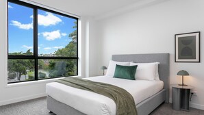 Un dormitorio moderno con un ventanal grande, una cama con cabecero gris, ropa de cama blanca y una manta verde.