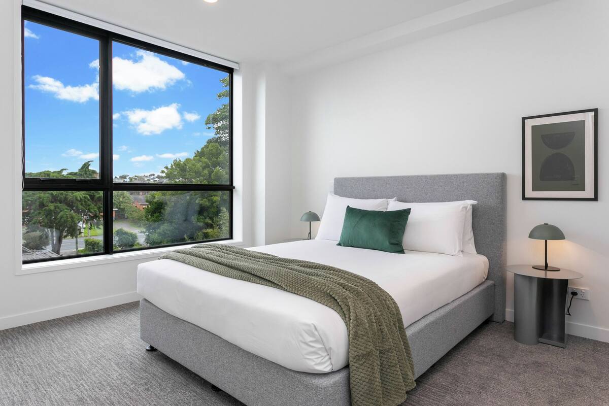 A modern bedroom with a large window, a bed with a grey headboard, white bedding, and a green throw blanket.