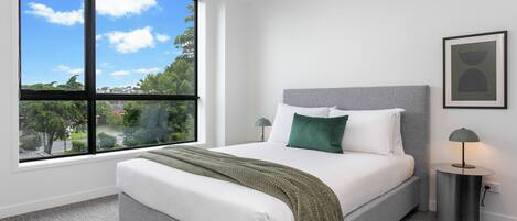 Une chambre moderne avec une grande fenêtre, un lit doté d’une tête de lit grise, une literie blanche et un plaid vert.