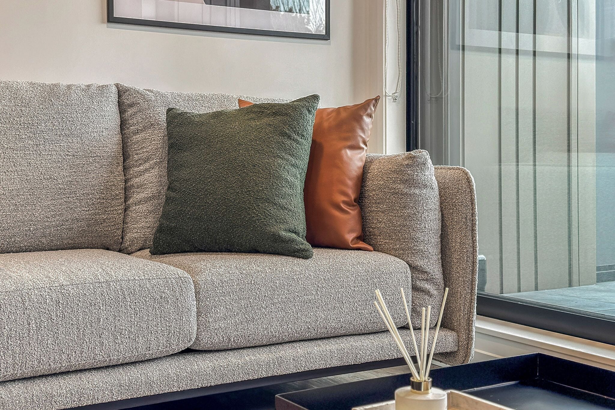 A grey sofa with a textured fabric, a green and a brown cushion, and a black tray with reed sticks.