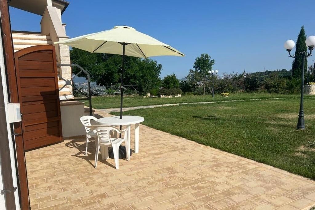 Een terras buiten met een tafel en stoelen onder een zonnescherm, een pad van bakstenen en een gazon met bomen.